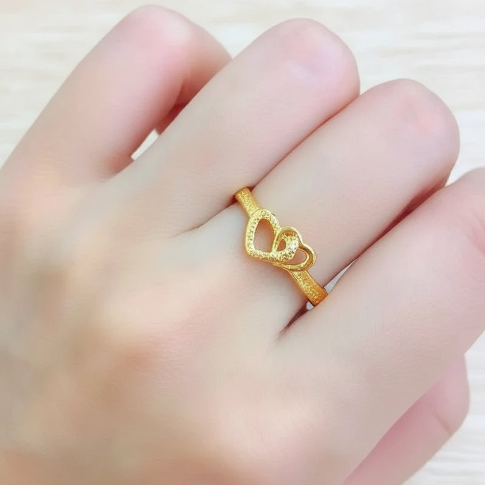 A person's hand wearing a gold-colored ring with a double interlocking heart design, featuring both textured and smooth polished finishes on the band and hearts.