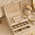 Beige jewelry box with jewelry inside on a table with flowers and a mirror.