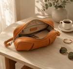 Brown leather bag on a table with a cup of coffee, sunglasses, and a plant in the background.