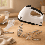 Hand mixer with attachments on a wooden surface, with eggs and a bowl in the background.