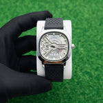 Watch with a black strap held by a hand on a white surface with grass in the background At Aezoon