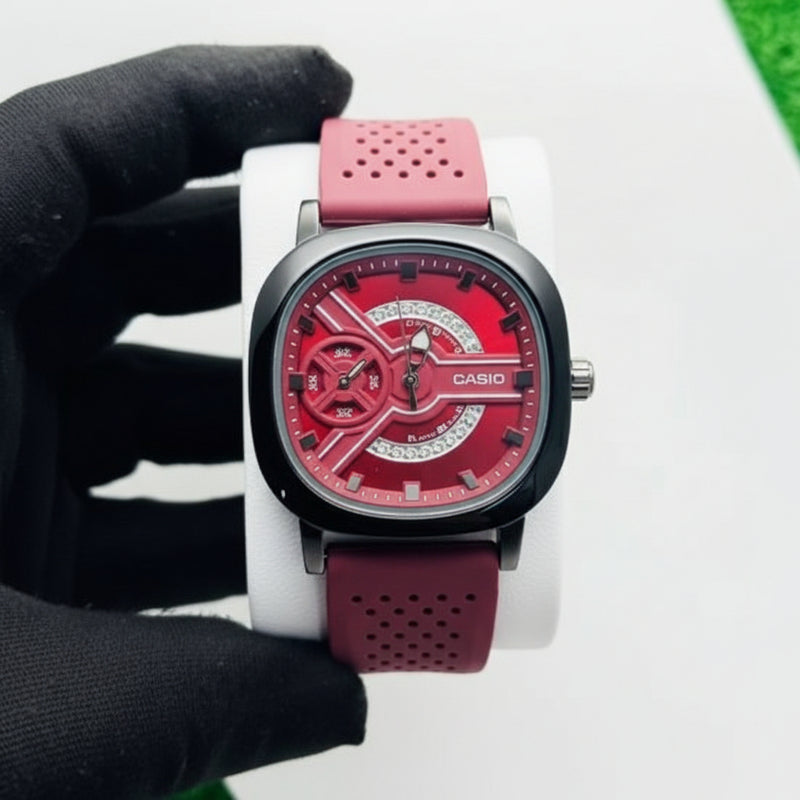 Watch with a red strap on a white surface, with a hand wearing a black glove holding it. At Aezoon